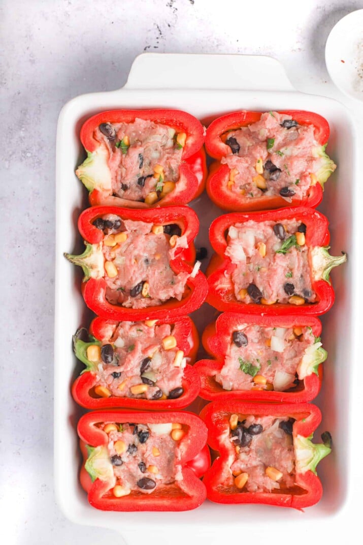 Eight filled red pepper halves in a white baking dish on a marble countertop