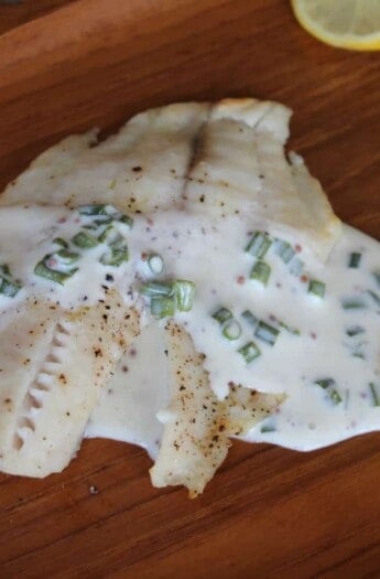A fillet of broiled tilapia covered in Mustard Chive Sauce next to a slice of lemon.