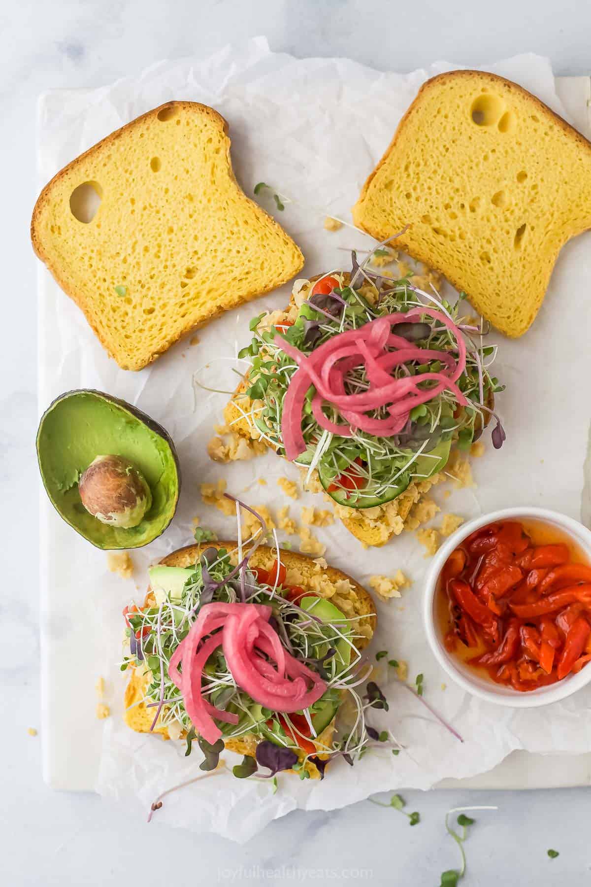 This Avocado Sandwich is the perfect quick lunch, with smashed chickpeas for protein, smoky roasted red peppers, and tangy pickled onions. Overhead view of two avocado sandwiches before adding second slice of bread