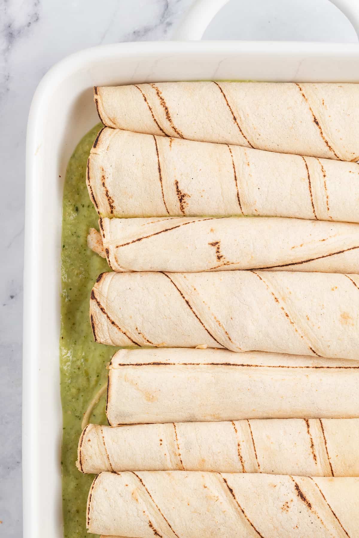 rolled tortillas in a white casserole dish on top of green sauce