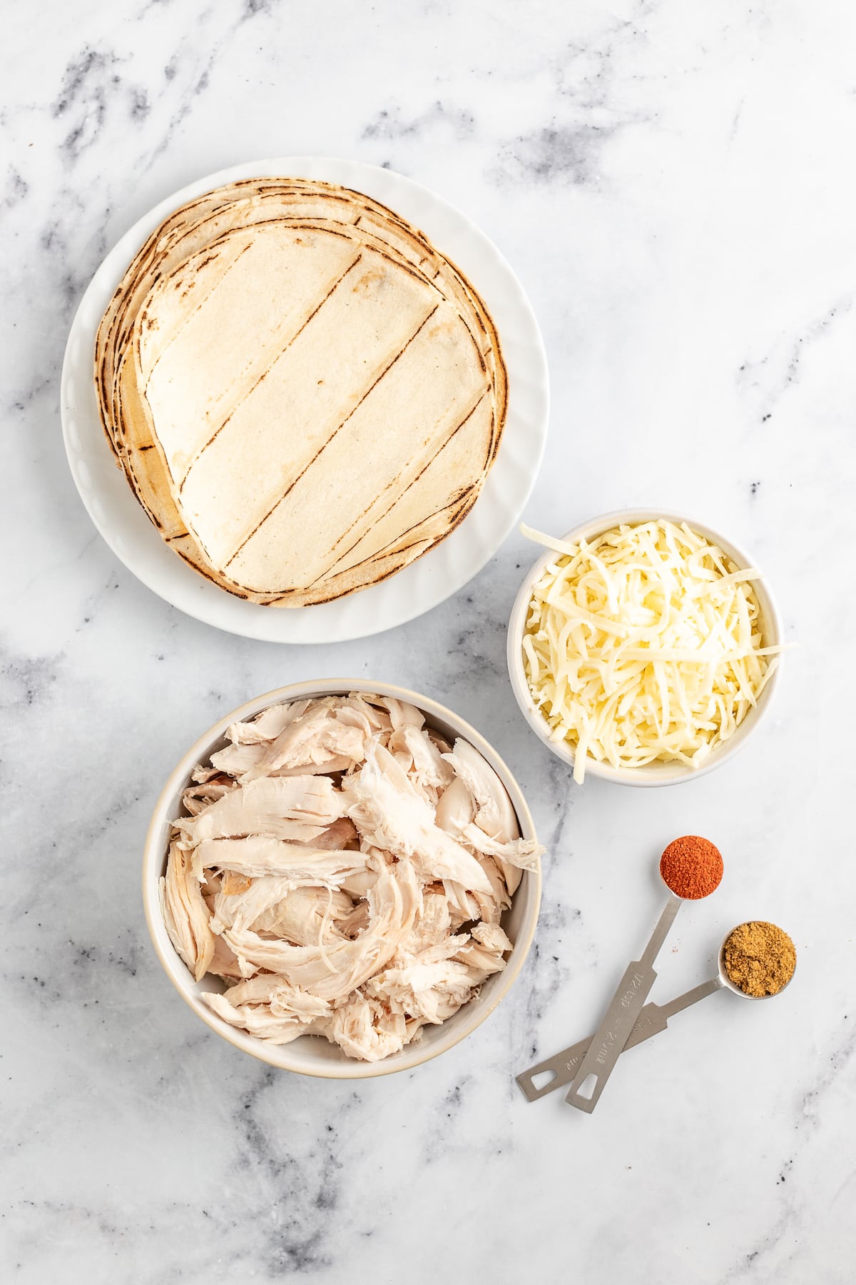 an assortment of tortillas, shredded cheese, chicken, and spices on a marble countertop