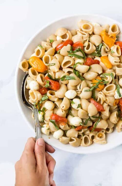 Roasted tomato caprese pasta salad in a bowl with a hand holding a spoon.