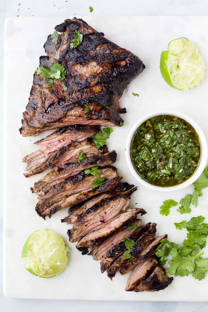 Grilled skirt steak on a cutting board with a bowl of chimichurri and lime