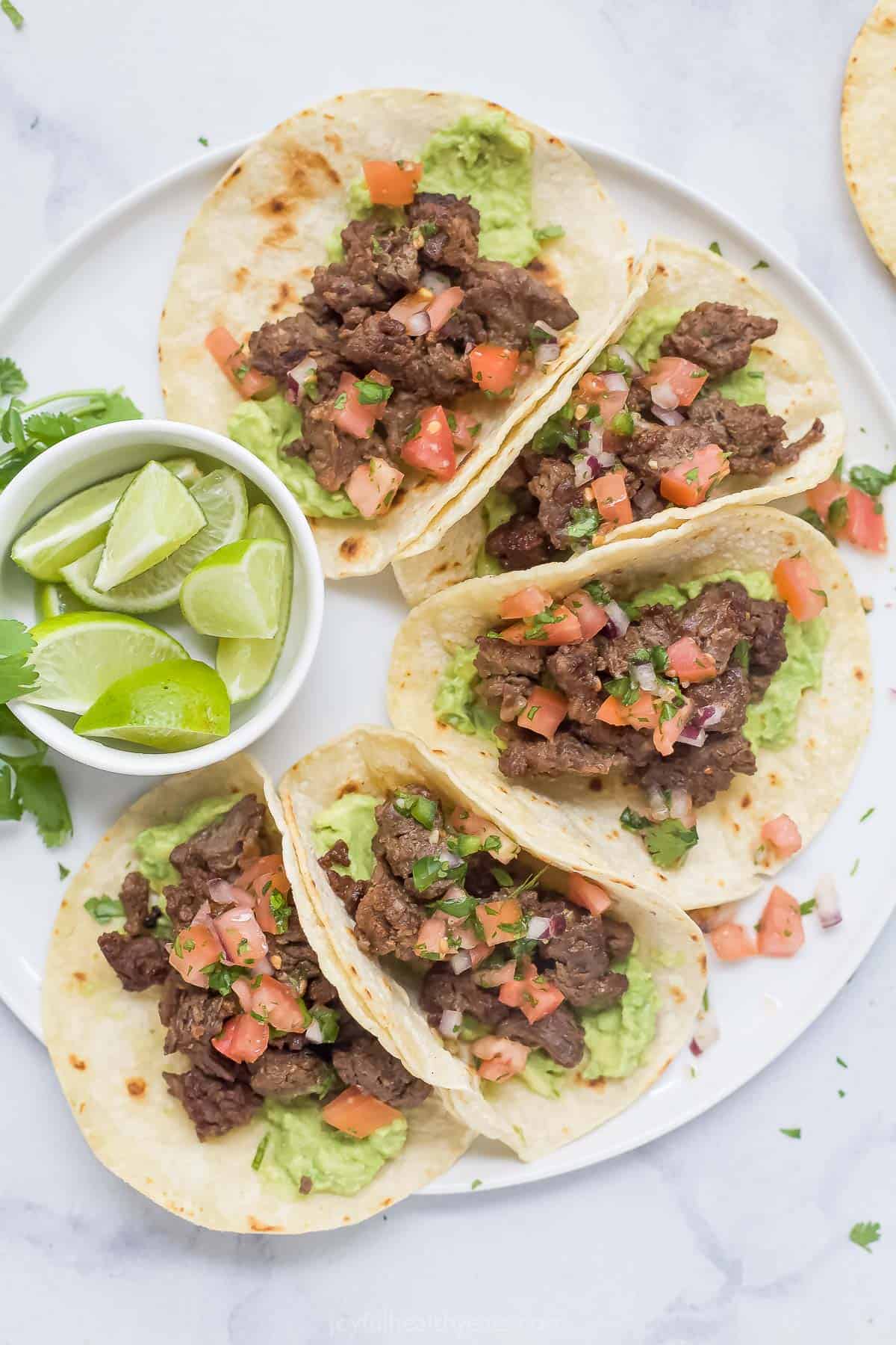 Plate of steak tacos with toppings and a bowl of lime wedges on the side. 