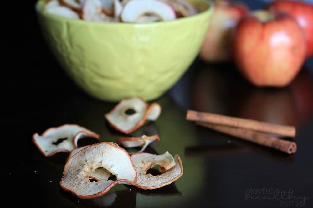 A few baked apple chips in front of a bowl of sliced apples and cinnamon sticks