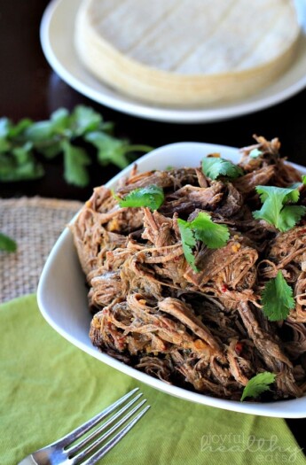 Crock Pot chipotle beef barbacoa inside of a bowl with a stack of tortillas on a plate behind it