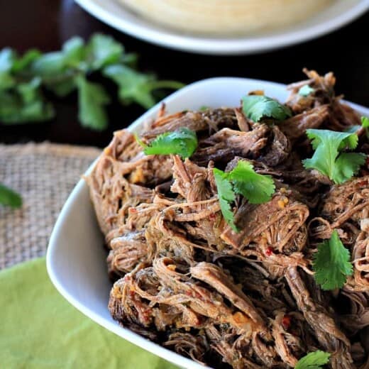 Crock Pot chipotle beef barbacoa inside of a bowl with a stack of tortillas on a plate behind it