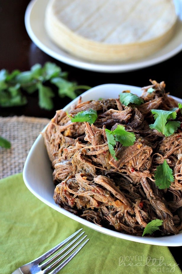 Crock Pot chipotle beef barbacoa inside of a bowl with a stack of tortillas on a plate behind it