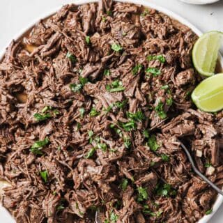 Bowl of chipotle crockpot barbacoa with fresh cilantro with lime wedges on the side.