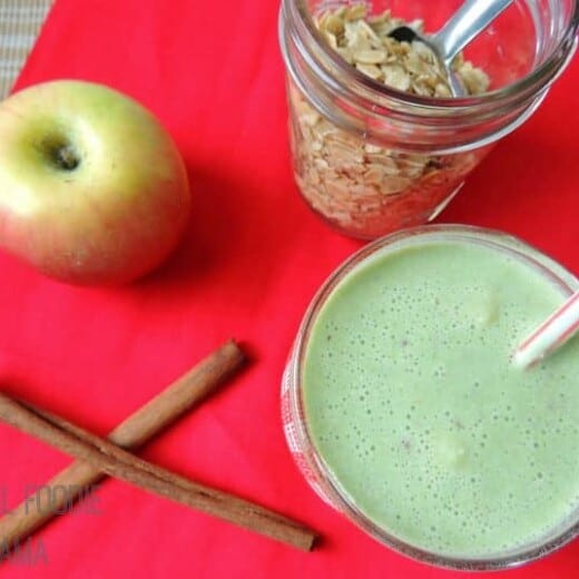 An Apple Ginger Crisp Smoothie Next to Two Cinnamon Sticks, an Apple and Granola