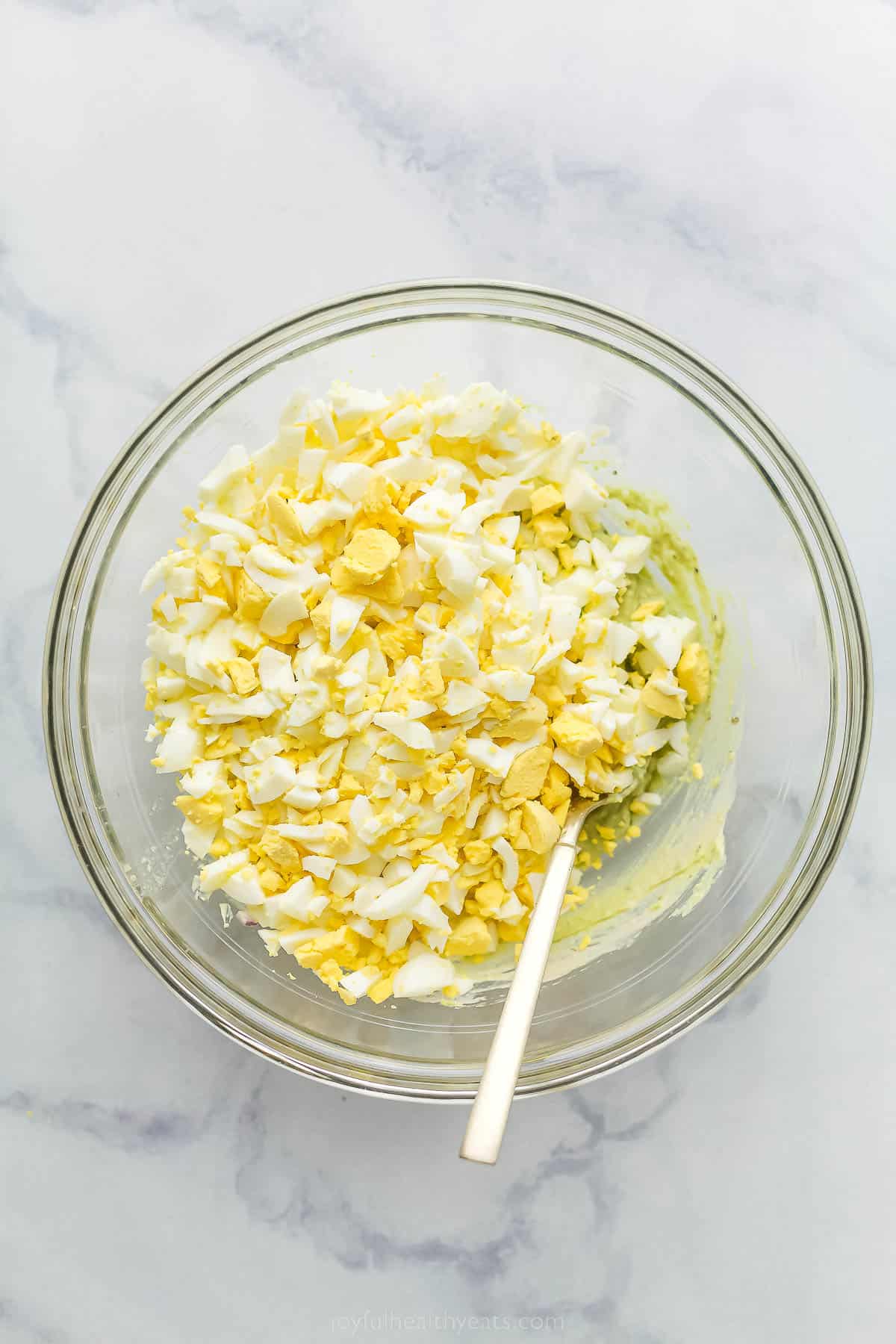 This Avocado Egg Salad is a healthy & satisfying salad made with less mayo! It's perfect for breakfast or lunch. a glass bowl with crumbled eggs and creamed avocado underneath