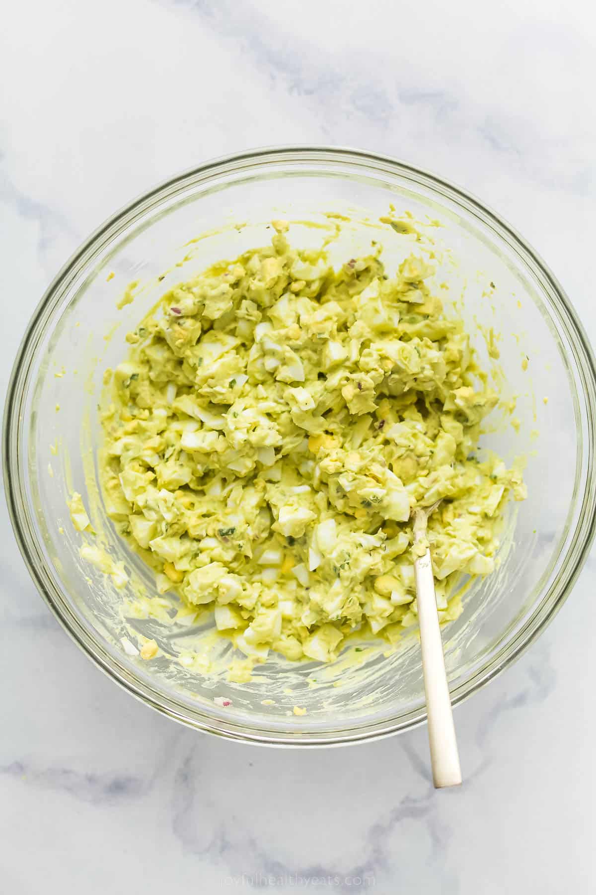 a glass bowl with egg salad being mixed with a wooden spoon