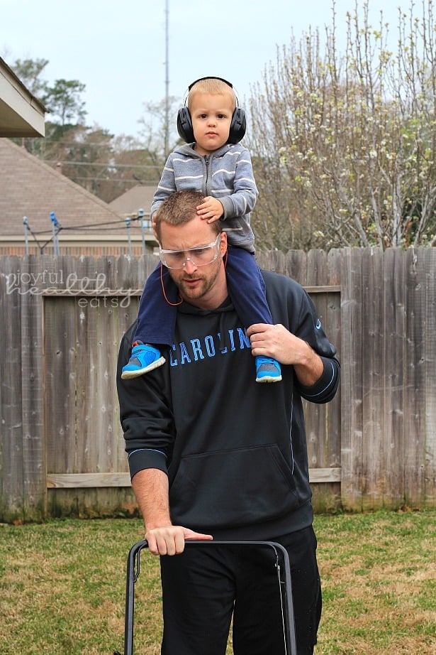 A toddler on his dad's shoulders while using a lawn mower