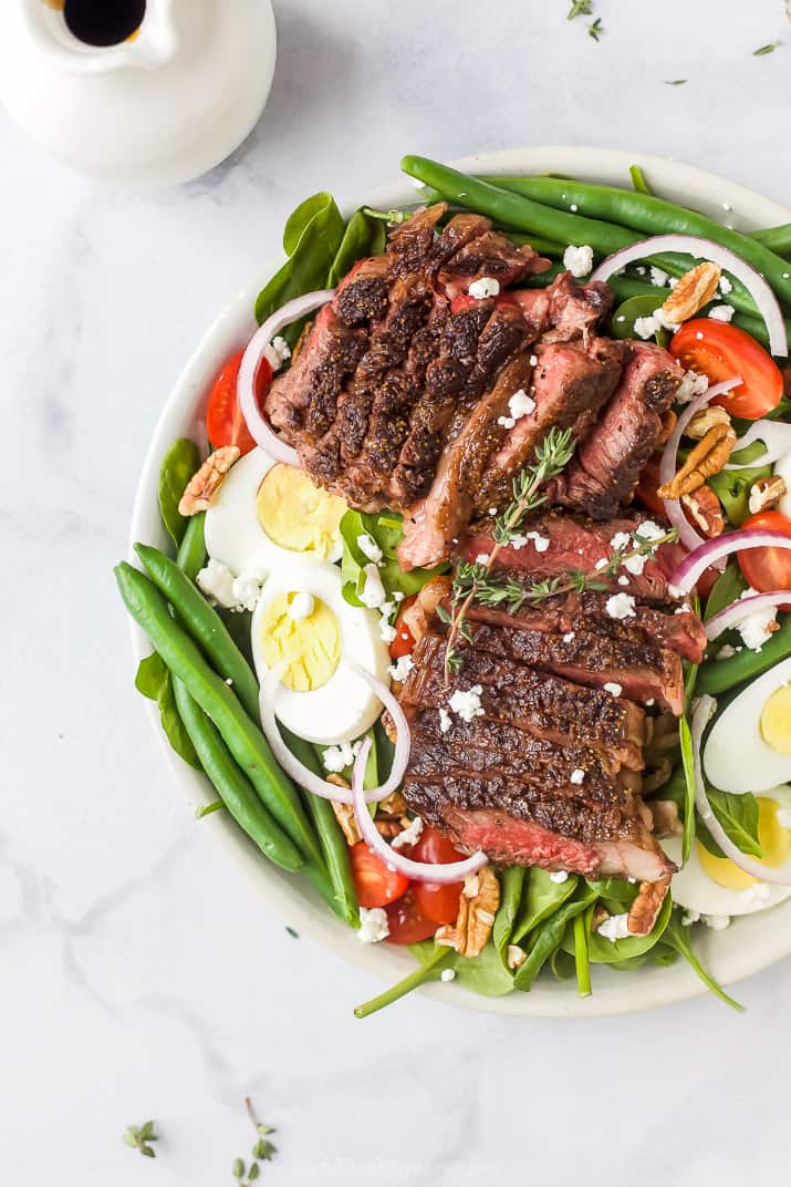 An overhead shot of a steak salad on a plate beside a container filled with balsamic vinaigrette