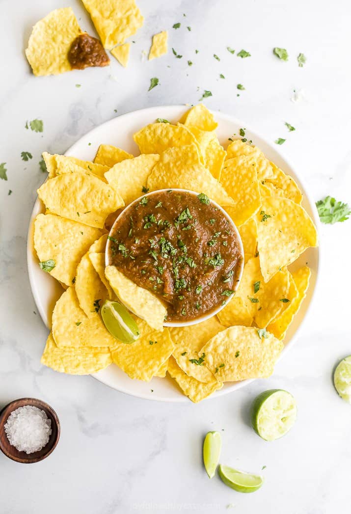 A round platter of tortilla chips with a smaller bowl of salsa in the center garnished with lime wedges and fresh cilantro