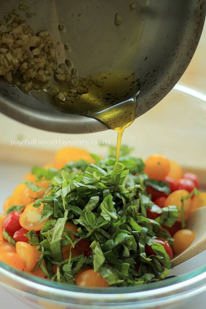 Garlic olive oil being poured over Bruschetta ingredients in a mixing bowl