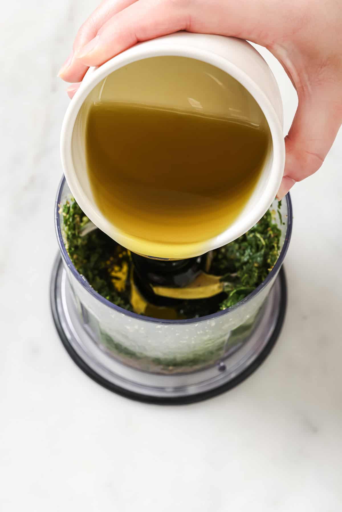 This Homemade Basil Pesto recipe is made with pecans instead of pine nuts to give it some extra body. It's my favorite way to make pesto! Pouring oil into food processor to make homemade basil pesto
