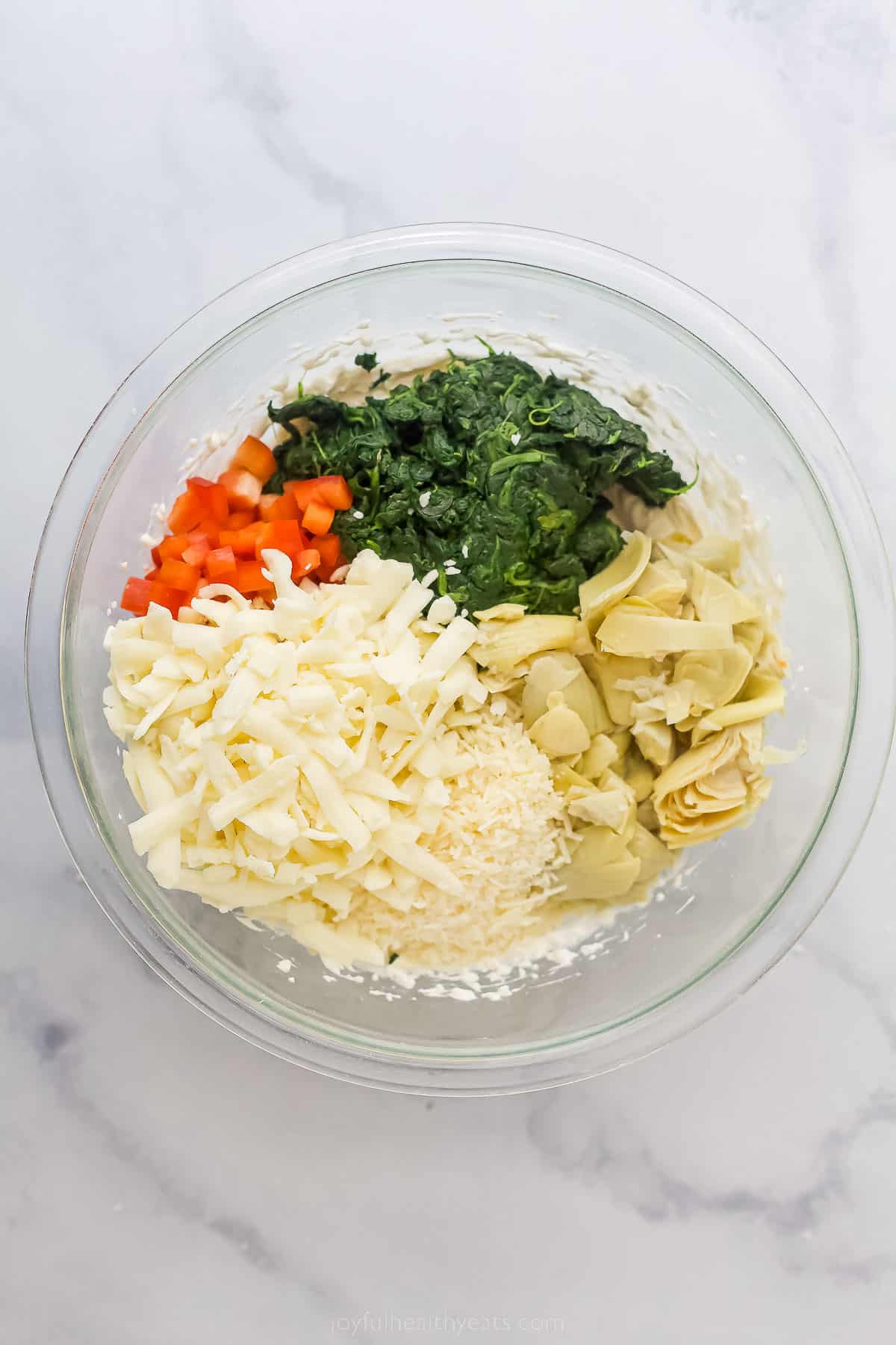 a glass bowl with artichokes, spinach, cheese, and red peppers