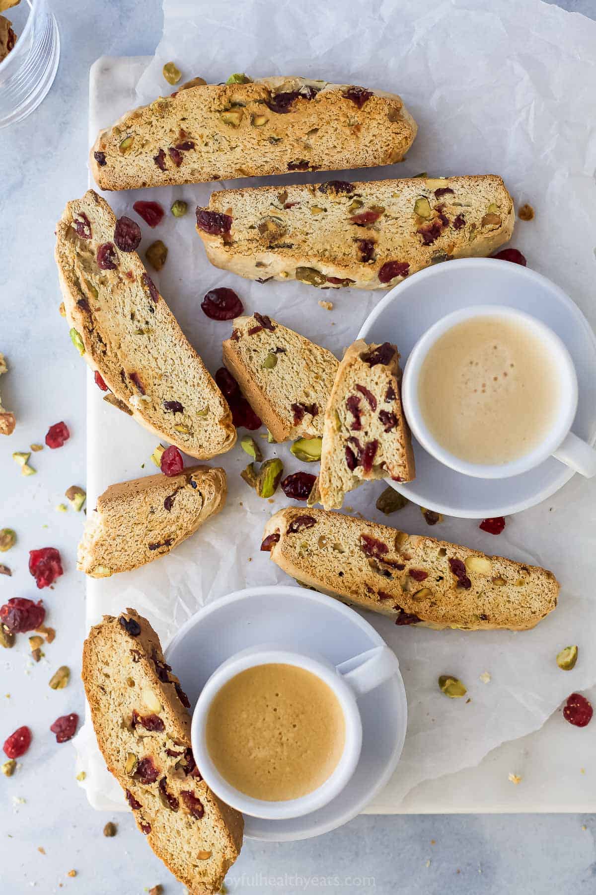 Two small mugs of coffee beside six pieces of cranberry pistachio biscotti on a countertop