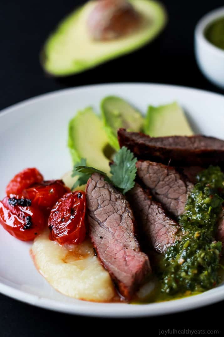 Close-up of Chili Rubbed Flank Steak slices on a plate served with fresh chimichurri sauce, roasted tomatoes, and fresh avocado