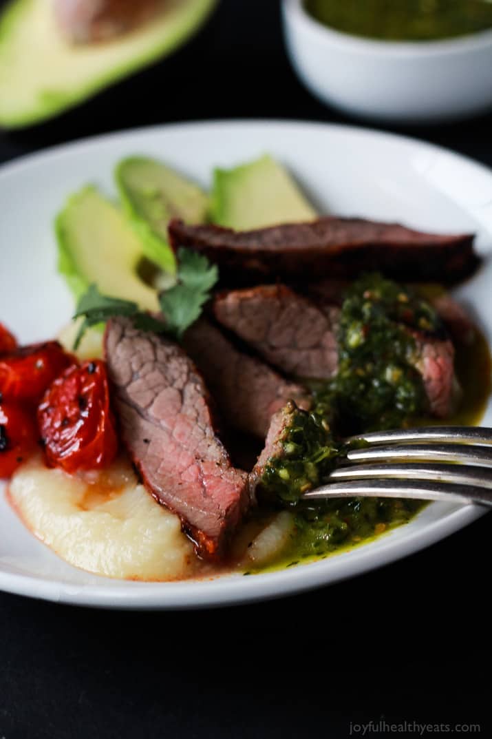 Close-up of slices of Chili Rubbed Flank Steak on a plate served with fresh chimichurri sauce, roasted tomatoes and fresh avocado