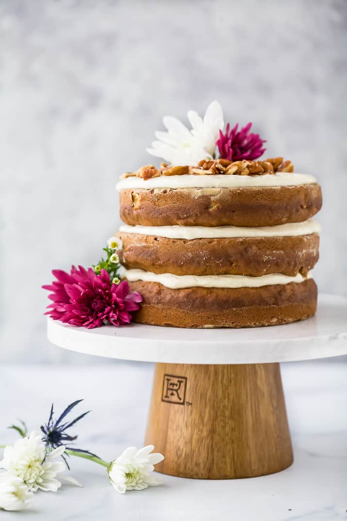 a three layer unfrosted cake with flowers as a garnish on a wooden and marble cake stand