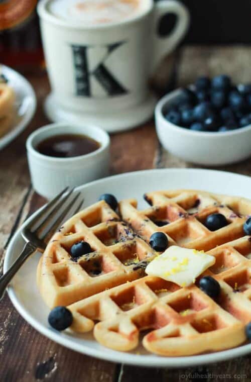 A Whole Wheat Lemon Blueberry Waffle on a Plate Next to a Coffee Mug and a Bowl of Blueberries