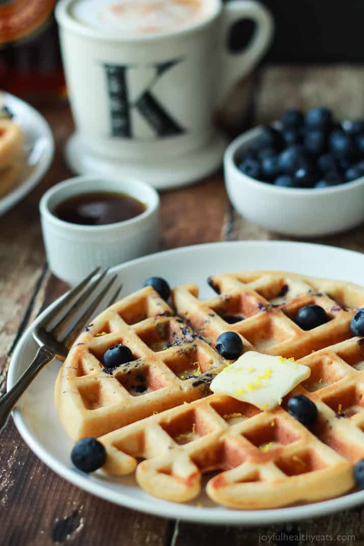 A Whole Wheat Lemon Blueberry Waffle on a Plate Next to a Coffee Mug and a Bowl of Blueberries