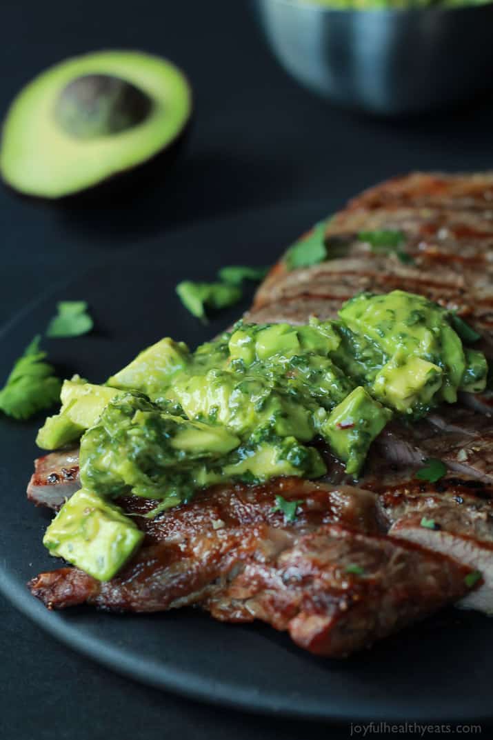 Close up of a Juicy Grilled Flank Steak topped with a fresh Avocado Chimichurri on a black plate