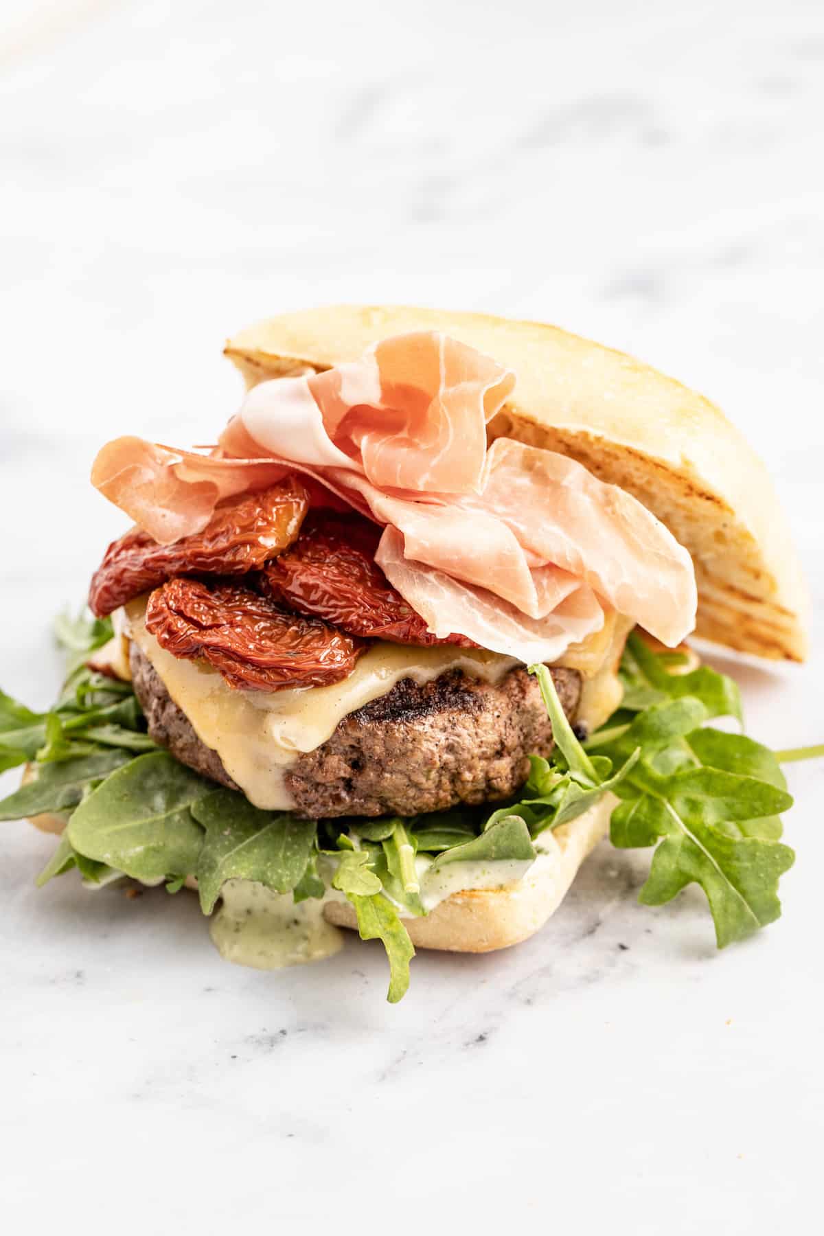 This 15-minute Italian cheeseburger is loaded with gouda cheese, sun-dried tomatoes, pesto aioli, and prosciutto for the perfect bite. Top bun resting on the side of the ciabatta burger.