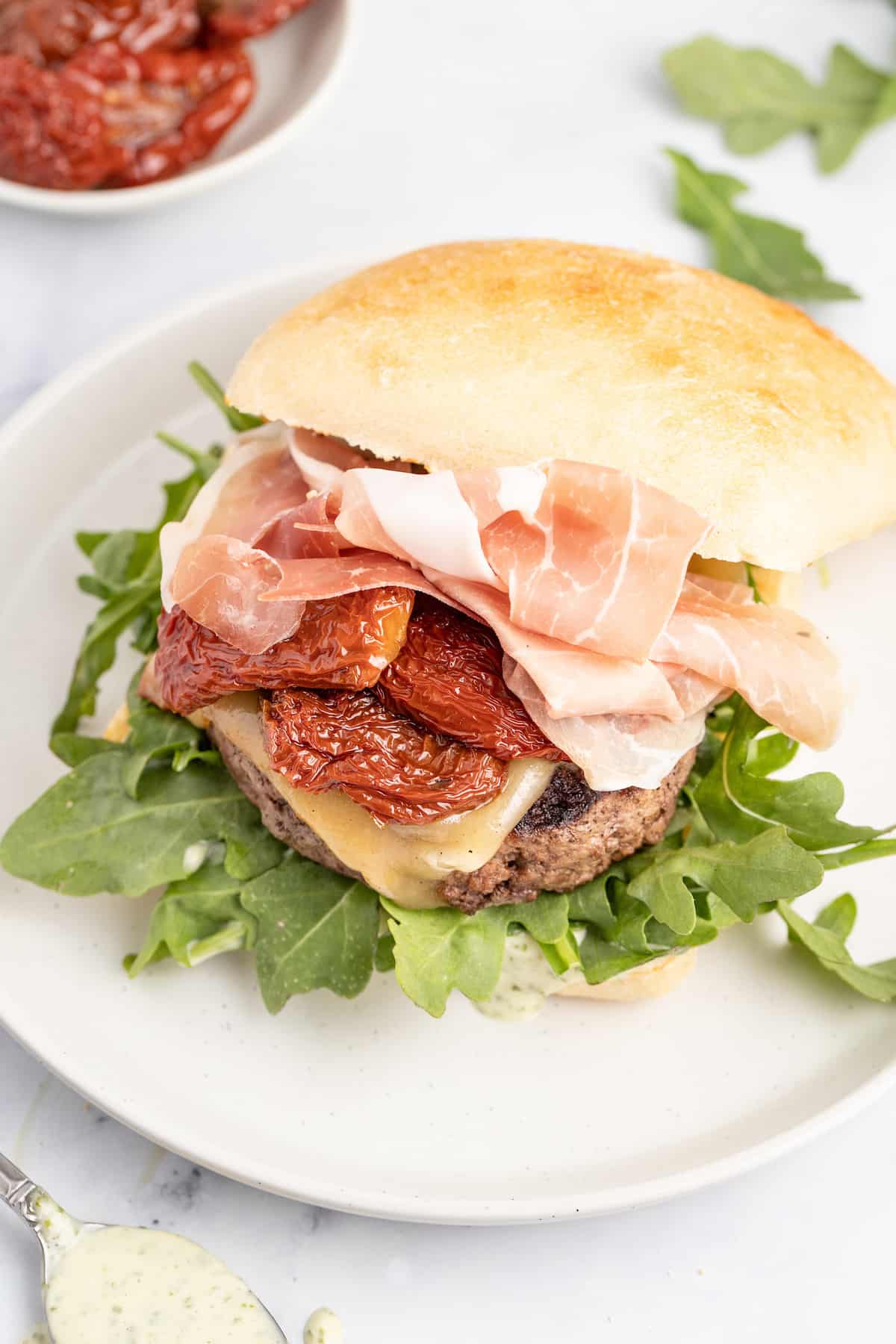 This 15-minute Italian cheeseburger is loaded with gouda cheese, sun-dried tomatoes, pesto aioli, and prosciutto for the perfect bite. Open Italian cheeseburger on a plate.