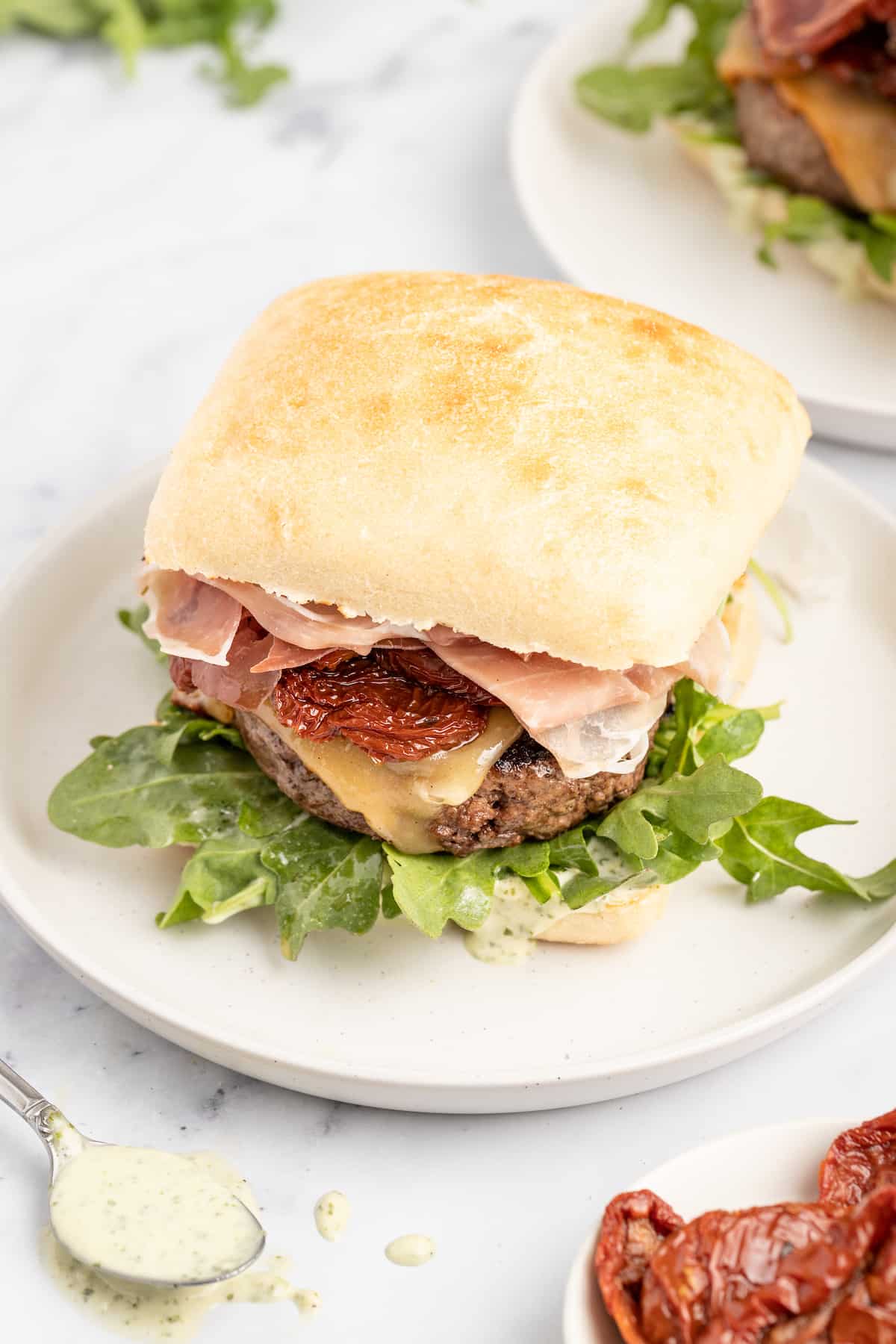 This 15-minute Italian cheeseburger is loaded with gouda cheese, sun-dried tomatoes, pesto aioli, and prosciutto for the perfect bite. Ciabatta burger on a plate.