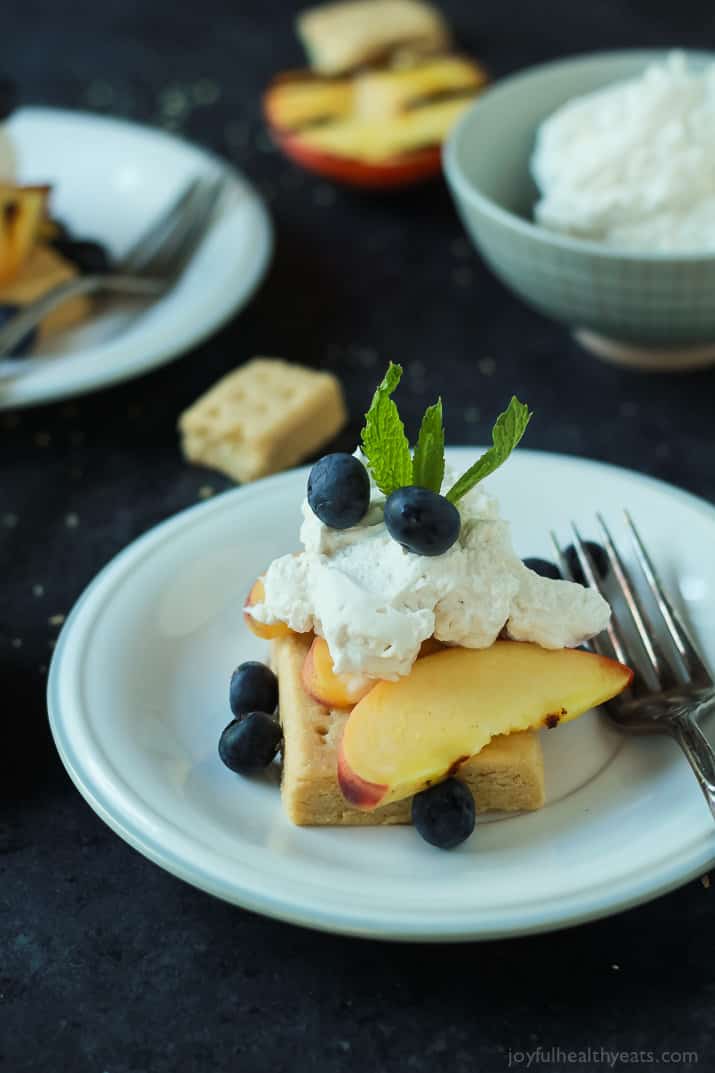 A serving of Grilled Peach Shortcake on a plate with shortbread, grilled peaches, coconut whipped cream, and fresh blueberries