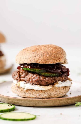 A lamb burger with whipped feta, caramelized onions, and sliced cucumbers on a serving plate.