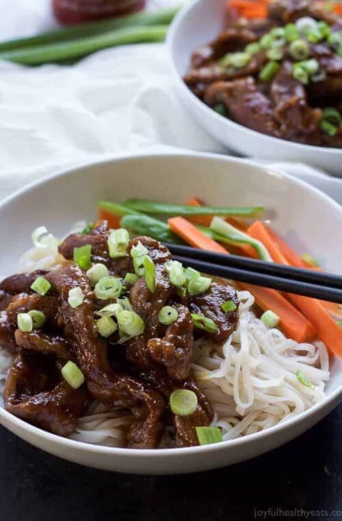 Sweet and spicy strips of flank steak in a shallow bowl with veggies, noodles and a pair of black chopsticks