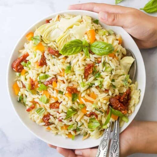 Two Hands Holding a Bowl of Mediterranean Salad with Two Spoons Inside