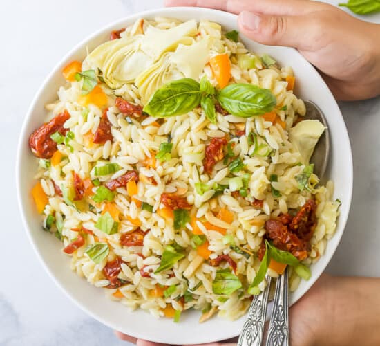 Two Hands Holding a Bowl of Mediterranean Salad with Two Spoons Inside