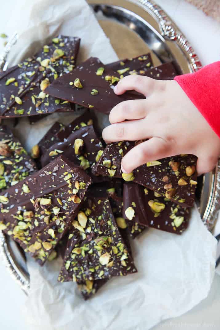 A child's hand reaching into a pile of Salted Pistachio Chocolate Bark on a platter