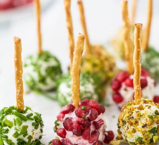 Nine assorted goat cheese balls on a sheet of parchment paper with a bowl of pomegranate seeds in the background