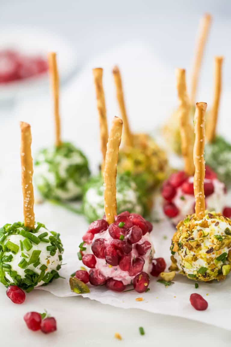 Nine assorted goat cheese balls on a sheet of parchment paper with a bowl of pomegranate seeds in the background
