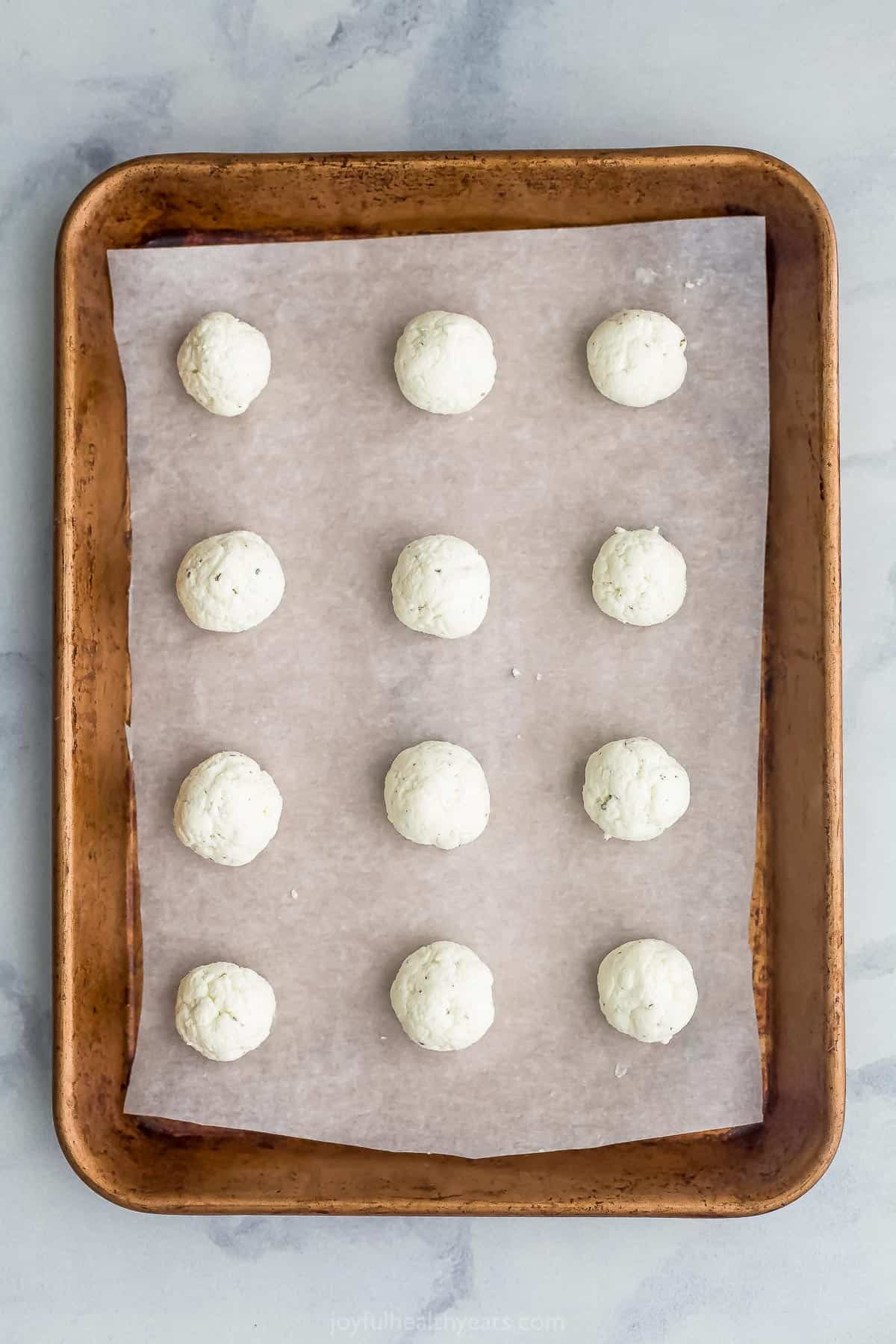 Twelve un-set cheese balls on a metal baking sheet lined with a piece of wax paper