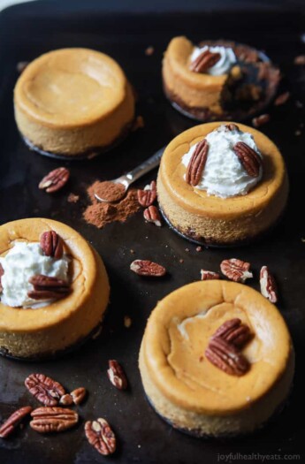 Five homemade single-serving Thanksgiving cheesecakes with pecans loose on the pan beside them
