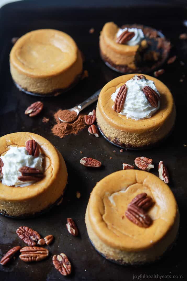 Five homemade single-serving Thanksgiving cheesecakes with pecans loose on the pan beside them