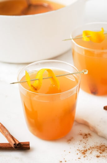 A glass of crockpot hot apple cider with an orange twist on a stirrer.