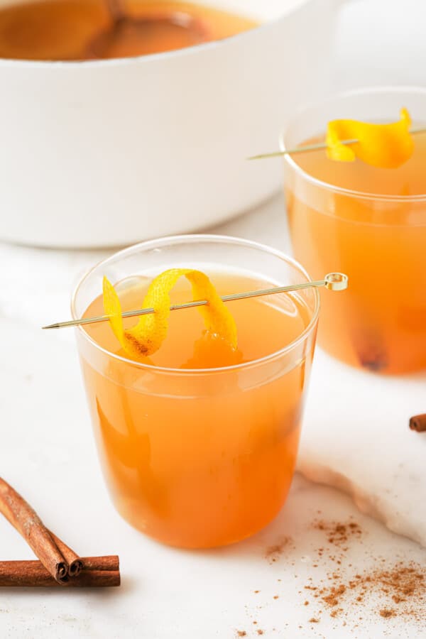 A glass of crockpot hot apple cider with an orange twist on a stirrer.