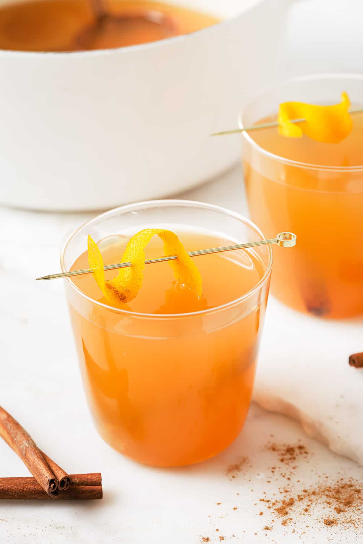 A glass of crockpot hot apple cider with an orange twist on a stirrer. 