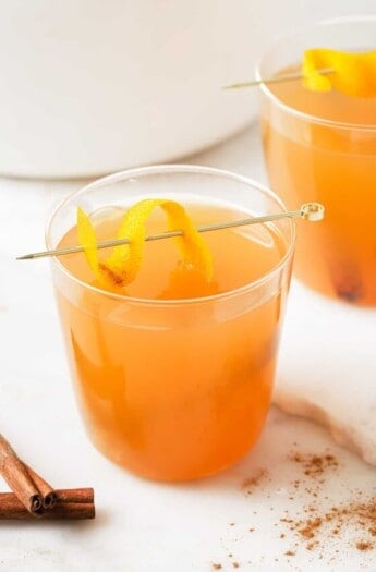 A glass of crockpot hot apple cider with an orange twist on a stirrer.