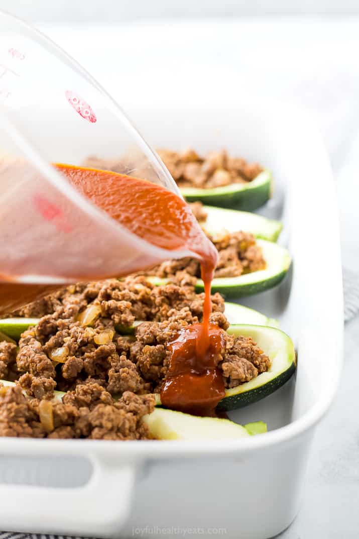 Red enchilada sauce being poured over zucchini boats stuffed with ground beef