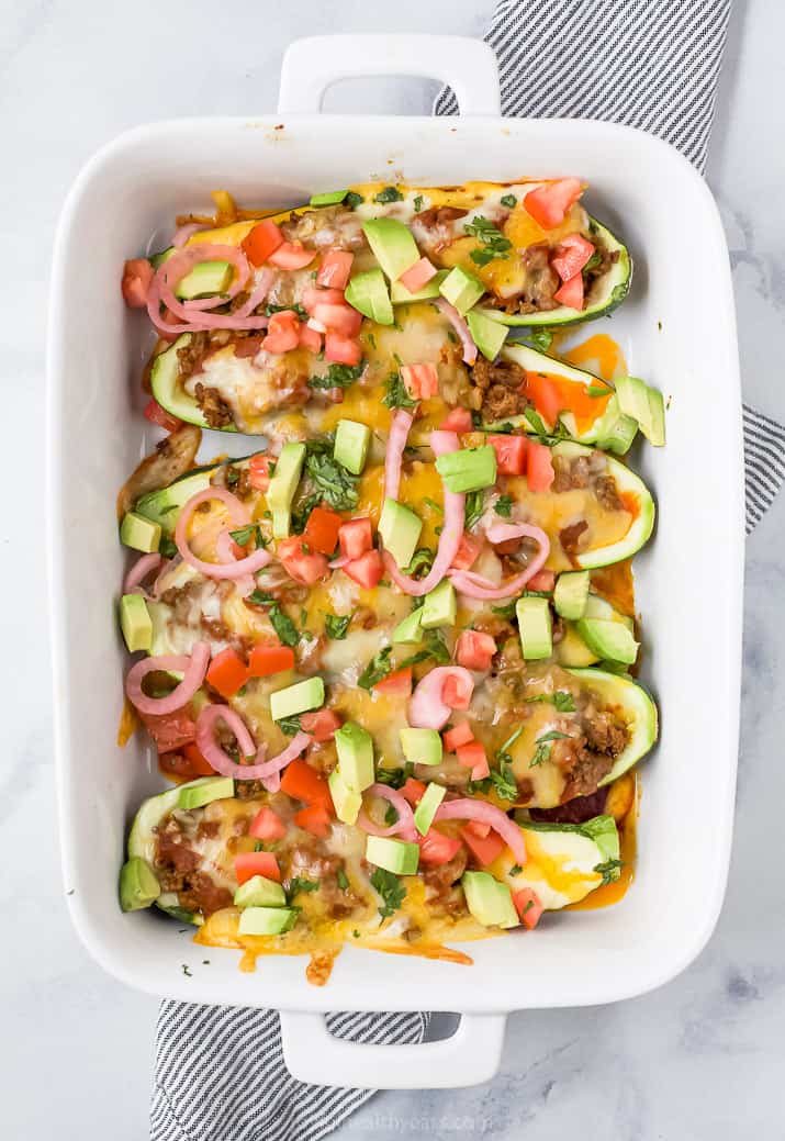 A white baking dish of stuffed zucchini boats topped with cheese, pickled onions, tomatoes and avocado