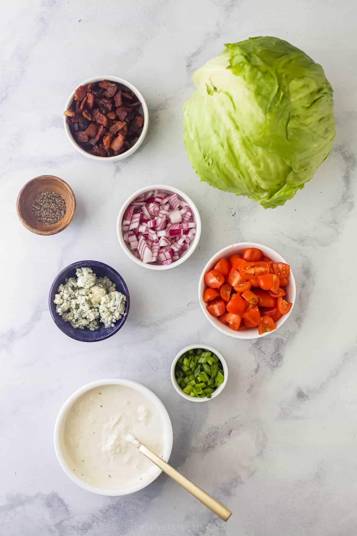 one head of iceberg lettuce and small bowls of bacon, red onions, tomatoes, green oniones, blue cheese, ground pepper, and blue cheese dressing